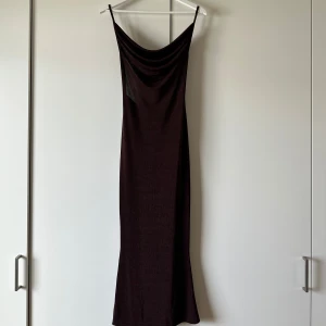 Brun långklänning med knyt i ryggen - Säljer en mörkbrun långklänning med smala axelband och snygg draperad urringning. Klänningen har en knyt-detalj i ryggen och är figurnära med ett mjukt, stretchigt material. Perfekt för dig som gillar minimalistisk och stilren look.