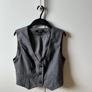 Grå väst från Gina Tricot - Snygg grå väst från Gina Tricot med djup v-ringning och tre knappar framtill. Västen har två fejkade fickor och är figursydd för en stilren look. Perfekt att styla över en skjorta eller t-shirt för en trendig vibe.