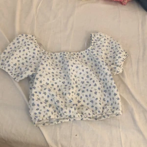 Blommig croppad topp från H&M - Supersöt croppad topp från H&M med vita och blå små blommor. Toppen har puffiga korta ärmar och resår vid hals och midja för en skön passform. Perfekt för varma dagar och ger en romantisk vibe till din outfit.