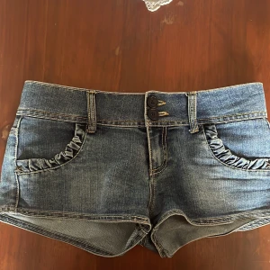 Blå jeansshorts med volangdetalj - Snygga blå jeansshorts med volangdetaljer på både fram- och bakfickor. Shortsen har två knappar och dragkedja framtill samt bälteshällor.  Väldigt låg midjade. Kontakta gärna innan köp. Köpta secondhand i England.