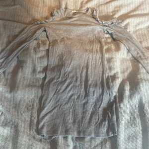 Grå ribbad klänning med volang - Grå ribbad kortklänning med lång ärm och snygga volangdetaljer vid axlarna. Klänningen har en offshoulder-design som ger en cool och trendig look. Perfekt för dig som gillar stilrena plagg med en twist.