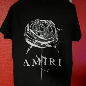 Svart Amiri t-shirt med tryck - Svart t-shirt från Amiri i bomull med coolt tryck på både fram- och baksida. Framsidan har Amiri-loggan med färgstänk som rinner ner, baksidan har en stor silverfärgad ros och Amiri-tryck. Klassisk passform och korta ärmar.