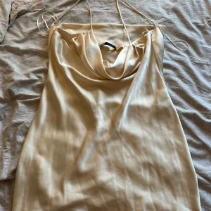 Champagnefärgad satin klänning från Zara - Champagnefärgad kortklänning från Zara i glansig satin med tunna axelband och snygg draperad urringning. Perfekt för dig som gillar minimalistisk och elegant stil. Klänningen är ärmlös och har en mjuk, följsam passform.