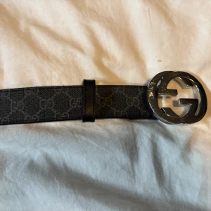 Svart Gucci bälte med GG-spänne - Säljer ett svart bälte från Gucci med klassiskt GG-mönster längs hela bandet och en stor, rund metallspänne med GG-logga i silver. Bältet är gjort i skinn och har en stilren, lyxig vibe som sticker ut.