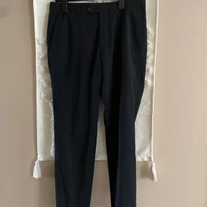 Svarta kostymbyxor Oscar Jacobson - Klassiska svarta kostymbyxor från Oscar Jacobson med rak passform och diskreta bakfickor. Byxorna har bälteshällor, knapp och dragkedja fram. Perfekta för en clean och stilren look.