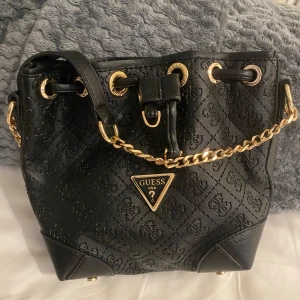Svart handväska från Guess med guldkedja - Snygg svart handväska från Guess med präglat logomönster och guldfärgade detaljer. Väskan har snörning upptill och en kedja som axelrem. Perfekt för dig som vill ha en trendig och stilren accessoar med lyxig känsla.