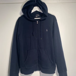 Polo Ralph Lauren Tech Performance Zip Hoodie  - Snygg marinblå hoodie från Polo Ralph Lauren med dragkedja, huva med snörning och klassisk logga på bröstet. Tech Performance modellen med nypris på 2100kr. Mjuk bomullskvalitet och känguruficka framtill. Perfekt för chill dagar eller när du vill ha en clean streetstyle. Välanvänd men i okej skick. 