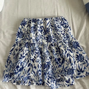 Vit och blå blommig kortkjol - Supersöt kortkjol med volanger i vitt och blått. Kjolen har en luftig och flowy passform, perfekt för varma dagar. 