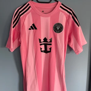 Inter Miami Messi 10 25/26 Matchtröja  - Inter Miami CF fotbollströja i rosa från Adidas, med svarta detaljer och klubbmärke på bröstet. Messi 10 tryck på ryggen. Tröjan har klassiska svarta Adidas-ränder på axlarna och är tillverkad i ett lätt, ventilerande material. Slim fit-modell med korta ärmar.