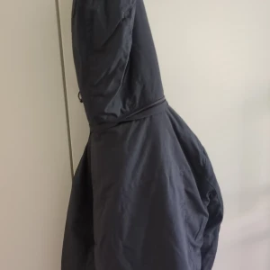 Svart parkajacka med huva och dragkedja - Säljer en svart parkajacka med huva, stora fickor med tryckknappar och dragkedja framtill. Jackan har även knappar som detalj och är perfekt för kyligare dagar. Materialet är bra  och jackan har en klassisk, lite längre passform.