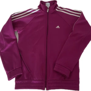 Adidas magentafärgad träningsjacka - Magenta träningsjacka från Adidas med vita ränder på axlarna och dragkedja framtill samt fickor på sidorna. Jackan har hög krage, långa ärmar och är tillverkad i mjuk polyester med Climawarm-foder. Perfekt för sport eller chill dagar. Det är tecken på användning då sliten.