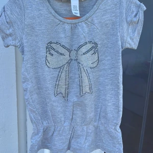 Grå topp med rosett från H&M - Supersöt grå topp från H&M med korta ärmar och en glittrig rosett framtill i silvriga stenar. Toppen har en liten volang nertill och är gjord i mjuk bomullsmix. Perfekt för dig som gillar lite extra bling på vardagen!