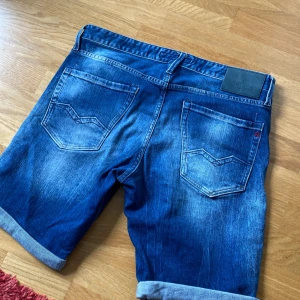 Blå jeansshorts från Replay - Snygga blå jeansshorts från Replay med klassisk femficksdesign och uppvikta benslut. Shortsen har en normal passform och coola slitningar för en avslappnad look. Perfekta för sommardagar när du vill ha en stilren men chill vibe.