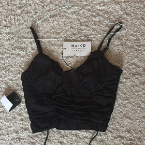Svart croppad korsettopp NA-KD - Supersnygg svart korsettopp från NA-KD med croppad passform och tunna justerbara axelband. Toppen har en unik skrynklig textur och snygg snörning framtill. Perfekt för att lyfta vilken outfit som helst med sin festliga stil.