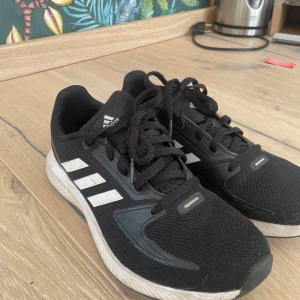 Svarta Adidas sneakers med vita detaljer - Svarta Adidas sneakers med klassiska vita ränder på sidorna och vit sula. Ovandelen är i mesh och syntet, vilket ger en sportig look och bra andningsförmåga. Skorna har snörning och rund tå, perfekt för dig som gillar en clean och stilren stil.
