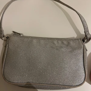 Glittrig silver axelväska - Säljer en snygg axelväska i glittrigt silver med dragkedja upptill. Väskan har en enkel, stilren design och ett axelband i samma glittriga material. Perfekt för att lyfta vilken outfit som helst med lite extra bling.