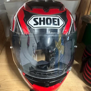 Shoei röd och svart motorcykelhjälm - Snygg integralhjälm från Shoei med röd, svart och silver design samt cool tribal-motiv på ovansidan. Hjälmen har klart visir och vadderad insida för extra komfort. Perfekt för dig som vill sticka ut på vägarna med en riktigt fet look.                                                                  Väldigt bra skick, har bara använts fåtal gånger.                Storlek: XS | 53-54 cm