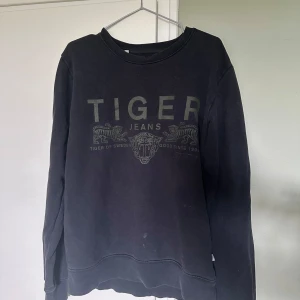 Svart sweatshirt från Tiger of Sweden - Svart sweatshirt från Tiger of Sweden med tryck framtill där det står 'TIGER JEANS' och två tigrar. Tröjan har rund halsringning och långa ärmar. Perfekt för en chill och stilren look.