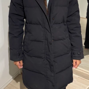 Svart lång dunjacka Canada Goose - Säljer en svart lång dunjacka från Canada Goose med huva och snygg pälskant. Jackan har raka linjer, knappar och dragkedja framtill samt logga på ärmen. Perfekt för kalla dagar när du vill hålla dig varm och ändå se stilren ut.