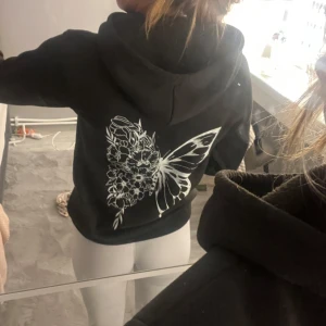 Svart hoodie med fjärilstryck - Svart hoodie med stor vit fjäril och blommor tryckt på ryggen. Klassisk huva med snörning och magficka framtill. Mjuk och skön, perfekt för chill dagar. Långärmad och i normal passform.