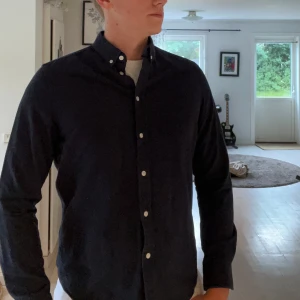 Mörkblå skjorta från Samsøe Samsøe - Stilren svart skjorta från Samsøe Samsøe med klassisk button down-krage och vita knappar. Skjortan har lång ärm och är tillverkad i mjuk bomull, perfekt för en clean och enkel look. Saknar storlekslapp men är i M