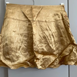 Guldig kjol  i sidenlook med knappar - Snygg guldig kjol med glansig finish och knappar i sidan. De har en lös passform och är tillverkade i ett material som ser ut som siden. Perfekta för dig som vill sticka ut med en lyxig vibe och gillar metallictoner. Tyvärr hinner jag inte att stryka den. Midjemått 68/70 cm