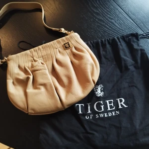 Beige axelväska från Tiger of Sweden - Säljer en beige axelväska i mjukt skinn från Tiger of Sweden. Väskan har veckad design framtill, justerbar axelrem och guldfärgade metalldetaljer. Invändigt finns dragkedjefack för småprylar. Perfekt för dig som gillar stilrena accessoarer.