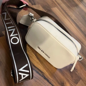  Väska Valentino - Säljer en stilren vit/beige axelväska från Valentino med bred brun axelrem med logga. Den är i nyskick och använd max 3 gånger. 