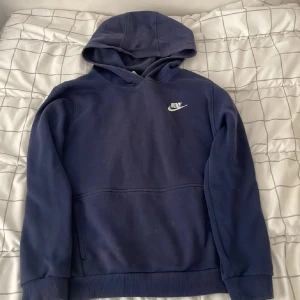 Mörkblå Nike hoodie XL - Säljer en mörkblå hoodie från Nike i storlek XL. Klassisk design med vit Nike-logga på bröstet, huva och känguruficka. Mjuk insida och ribbade muddar vid ärmslut och nederkant. Perfekt för chill dagar eller när du vill ha en sportig look.