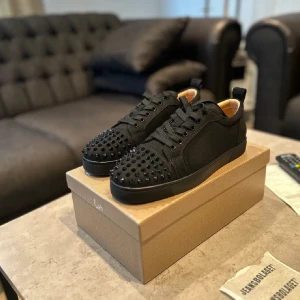 Svarta Louboutin sneakers med nitar - Snygga svarta sneakers från Louboutin med mattsvarta nitar på tån och den ikoniska röda sulan. Skorna har snörning, rund tå och är tillverkade i textil och skinn. Perfekta för dig som vill sticka ut med en edgy och lyxig stil.