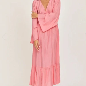 Rosa långklänning med v-ringning - Superfin rosa långklänning med v-ringning och vida ärmar. Klänningen har knappar framtill, volang nertill och öppen rygg med knytband. Tillverkad i ett lätt, krinklat tyg som känns luftigt och perfekt för varma dagar. Använd 1 gång. Nypris 699kr ‼️Lånade bilder