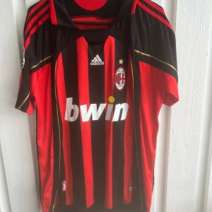 AC Milan Kaka' 22 Adidas fotbollströja - Säljer en klassisk AC Milan fotbollströja från Adidas med Kaka' och nummer 22 på ryggen. Tröjan är röd och svart med guldiga detaljer, randigt mönster och klubbmärke på bröstet. Kortärmad modell i lätt och ventilerande material, perfekt för fotbollsälskare.