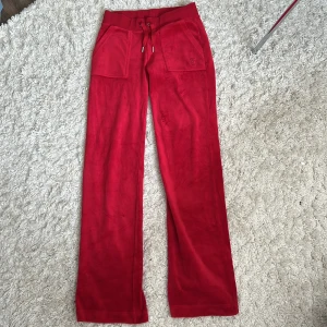 Röda Juicy Couture XS - Röda mjukisbyxor från Juicy Couture i velour med bred resår och snörning i midjan. Byxorna har raka ben, stora fickor fram och bak samt broderad JC-logga på ena fickan. Perfekta för en chill och trendig look. Använt 1/2 gånger 