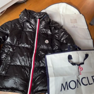 Svart pufferjacka från Moncler - Snygg svart pufferjacka från Moncler med glansig finish och klassisk logga på bröstet. Jackan har dragkedja med röd, vit och blå rand, hög krage och praktiska fickor. Perfekt för kalla dagar när du vill hålla stilen. Boxen med oxå nfc tagen funkar 