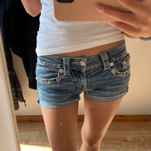 Miss Me jeansshorts med detaljer - Jättesöta blå jeansshorts från Miss Me med broderade detaljer och bling på fickorna. Shortsen har uppvikta benslut och dekorativa knappar. Shorten är ganska små i storleken så skulle snarare säga att de är i stolek 23. 
