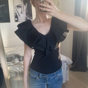 Svart volangtopp med v-ringning - Säljer en svart topp med v-ringning och stora volanger över axlarna. Toppen är figurnära och har en riktigt snygg och trendig look som sticker ut. Perfekt för dig som gillar statement-plagg och vill ha något unikt i garderoben. Var från början en klänning som jag klippt till topp! Aldrig använd, dragkedja i ryggen💞