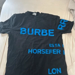Svart Burberry t-shirt med blå text - Cool svart t-shirt från Burberry med stora blå tryckta bokstäver och textdetaljer över hela plagget. Låg precis då den är använd men är väldigt fräsch och garanterar dig blickar runt om