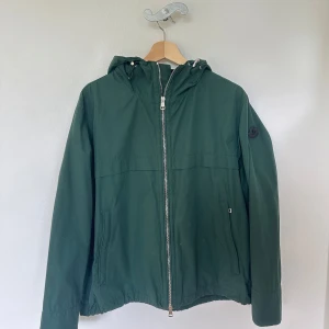 Grön vindjacka från Moncler  - Snygg grön vindjacka från Moncler med huva och dragkedja framtill. Jackan har Moncler-logga på ärmen, två fickor med dragkedja och meshfoder på insidan.  Köptes som present till min pojkvän i slutet av 2024 på Vestiare Collective men då den var för liten för honom säljer jag den nu vidare. En modell som är svår att få tag på idag.  Skriv för mer info! Inga defekter finns. Storlek 2. Modellen heter Yohann Giubbotto. Finns kvitto från Vestiare. 