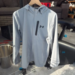 Ljusgrå vindjacka från Under Armour - HELT NY OCH ÄKTA Snygg ljusgrå vindjacka från Under Armour med huva och svart logga på bröstet. Jackan har en bröstficka med dragkedja och är tillverkad i ett lätt syntetmaterial. Perfekt för träning utomhus eller när du vill ha en sportig look. Fick den som present men den va för stor så det är därför jag säljer den 