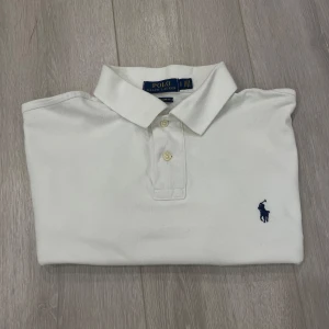 Vit Piké från Polo Ralph Lauren - Product Description: Selling this authentic Polo Ralph Lauren short-sleeve polo shirt in a crisp white color. Size M (Men's). In excellent condition - gently worn, no stains or flaws. Made from 100% cotton, this premium piqué fabric is soft, breathable, and high quality. Features the iconic navy embroidered Polo logo on the chest and a classic two-button collar. Style Tip: Dress it up with chinos or dress it down with jeans or shorts. Perfect for casual days, smart-casual events, or layering und