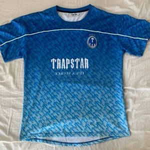 Blå fotbollströja Trapstar 22 - Snygg blå fotbollströja från Trapstar med stort tryck på bröstet och ryggen. Tröjan har ett mönster över hela ytan, vita detaljer och nummer 22 på ryggen. Tillverkad i lätt och ventilerande material, perfekt för fotbollsplanen eller streetwear.