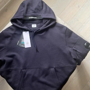 Mörkblå hoodie från C.P. Company XL - Snygg mörkblå hoodie från C.P. Company i fleece med huva och snörning. Tröjan har en ikonisk linsdetalj på ärmen och en praktisk ficka med dragkedja. Perfekt för dig som gillar streetwear och vill ha något bekvämt och stilrent. Aldrig använd med prislapp kvar, skick 10/10. Nypris 2899kr, XL fits L!