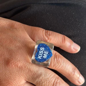 Statementring med blått hjärta st16,9 - Cool statementring i transparent plast med ett blått glittrigt hjärta på ovansidan där det står 'KISS ME' i vitt. Ringen har en chunky, fyrkantig form som verkligen sticker ut och ger en lekfull vibe till din stil.