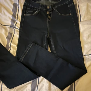 Mörkblå jeans från Blooming med hjärtknappar - Mörkblå jeans från Blooming med kontrastsömmar, hjärtformade knappar och broderade hjärtan på bakfickorna. Klassisk femficksmodell i jeansmaterial och normal passform. Perfekta för dig som gillar unika detaljer och snygg stil.