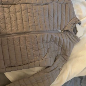 Beige dunjacka från Moncler - Säljer en beige dunjacka från Moncler med klassisk quiltad design och dragkedja framtill. Jackan har Moncler-logga på ärmen och står för både stil och funktion. Perfekt för kyliga dagar och enkel att matcha med olika outfits.