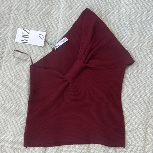 Vinröd oneshoulder topp från Zara - Säljer en vinröd ribbad oneshoulder topp från Zara med en stor rosettdetalj framtill. Toppen har en croppad passform och är tillverkad i ett stretchigt material som sitter snyggt. 