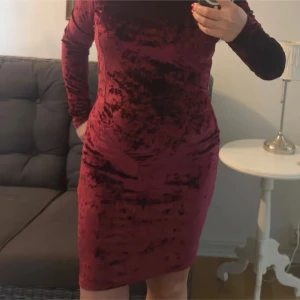 Vinröd sammetsklänning med lång ärm - Supersnygg vinröd sammetsklänning med lång ärm och tight passform. Klänningen är kort och har en rund halsringning. Materialet är mjukt och glansigt med en lyxig känsla. Perfekt för dig som vill sticka ut med en elegant look.