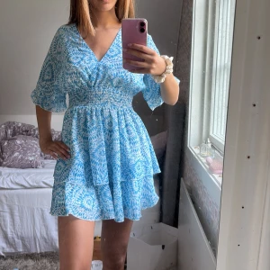 Blåvit kortklänning - Supersöt blåvit kortklänning med v-ringning, smockad midja och volangig kjol. Klänningen har ett blommigt mönster, vida korta ärmar och är gjord i ett lätt material som passar perfekt till varma dagar.