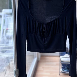 Svart croppad långärmad topp med knyt - Snygg svart croppad topp med långa ärmar och djup rundad urringning framtill. Toppen har en söt knytdetalj vid bysten och lätt rynkade partier som ger en trendig look. 