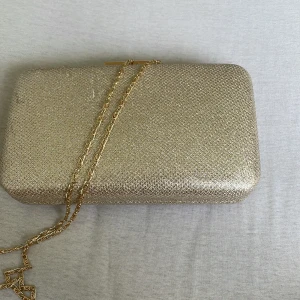Glittrig guldig kuvertväska med kedja - Säljer en fyrkantig kuvertväska i glittrigt guld med snygg guldfärgad kedja. Väskan har hårt skal och stängs med ett elegant guldfärgat spänne. Perfekt för att lyfta din outfit med lite extra bling.